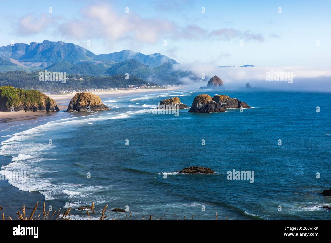 Vista panoramica verso Cannon Beach e Haystack Rock in una giornata di sole in Oregon. Dietro Haystack Rock arriva un muro di nebbia. Foto Stock