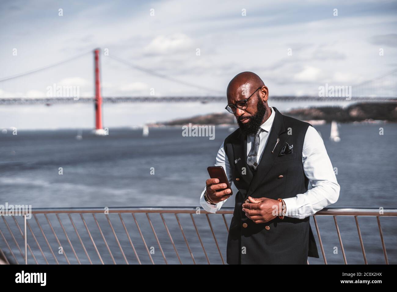 Un imprenditore africano serio ed elegante calvo in costume e occhiali sta usando il suo smartphone mentre si appoggia contro la scherma con un fiume Foto Stock