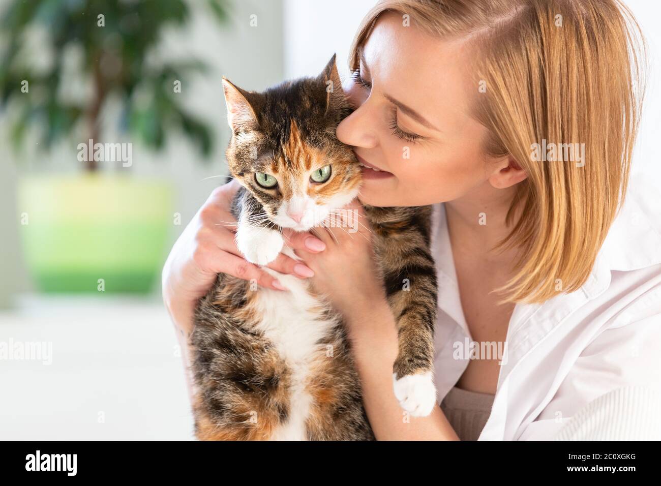 Primo piano di donna sorridente in camicia bianca baciando, abbracciando, abbracciando con tenerezza e amore gatto domestico in casa. Amore per gli animali, concetto di animali domestici Foto Stock