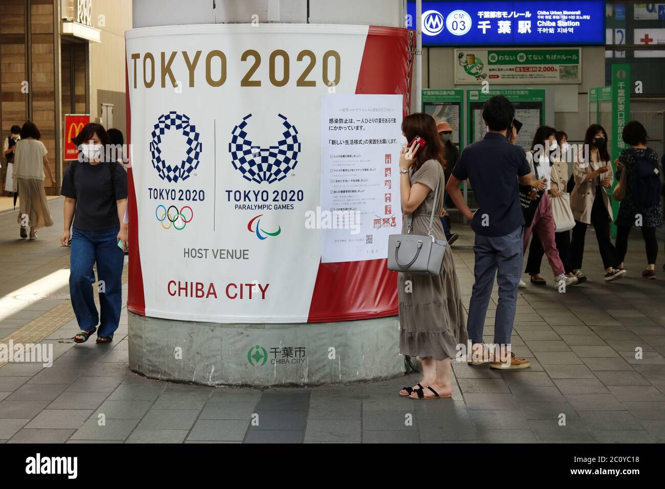 Una colonna avvolta di fronte alla stazione di Chiba con il logo delle Olimpiadi di Tokyo e un poster con consigli sulle misure di sicurezza dei coronavirus. Le persone indossano maschere facciali. Foto Stock