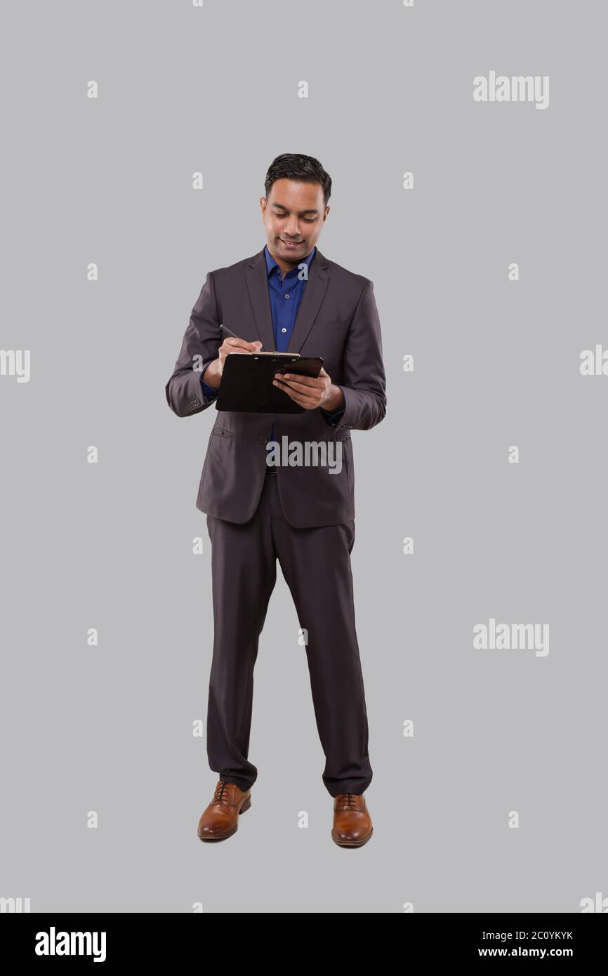 Uomo d'affari che scrive in Clipboard isolato. Uomo d'affari indiano in piedi a lunghezza intera con le clipboard in mani Foto Stock
