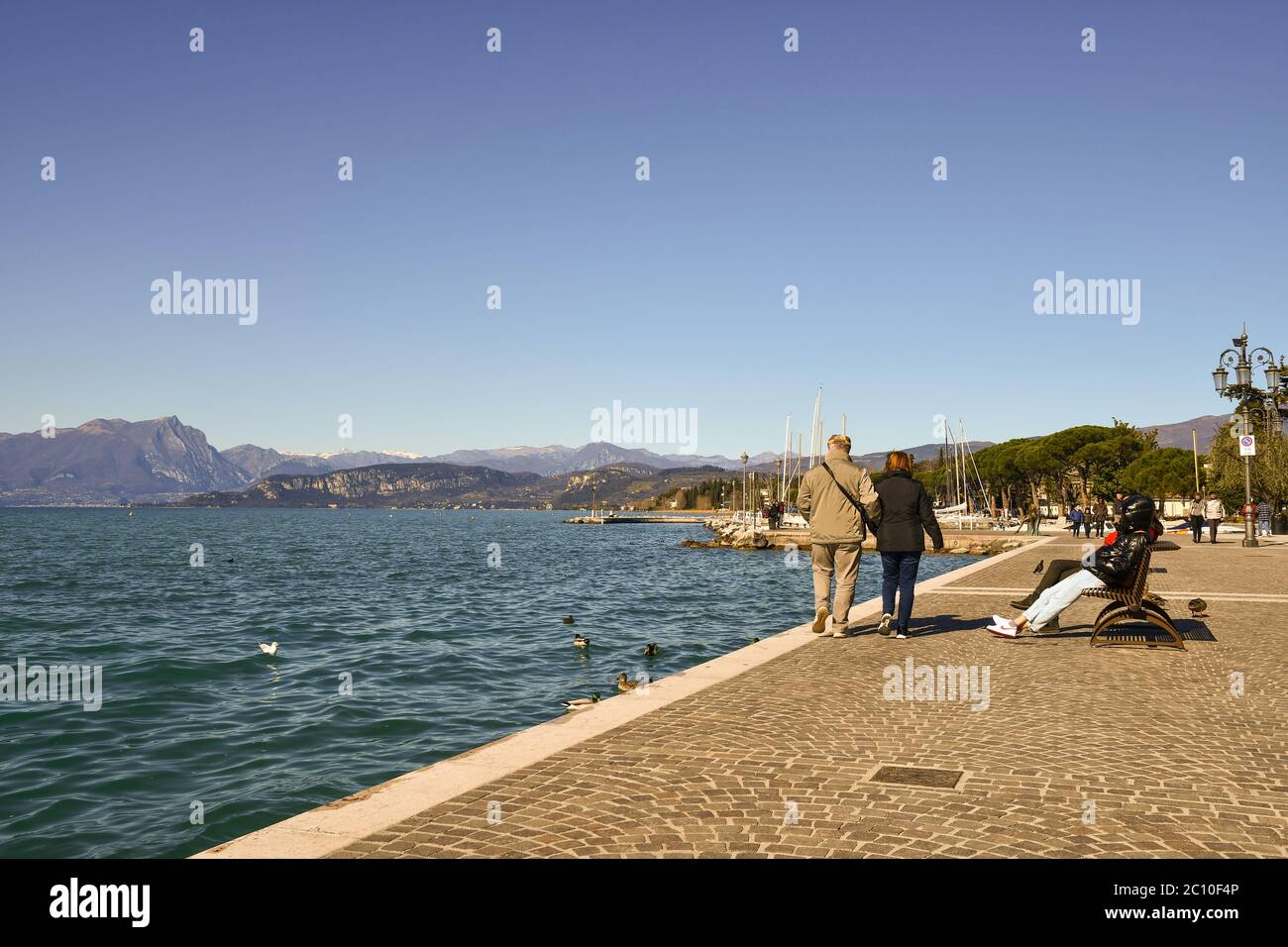 Vista panoramica sul Lago di Garda con persone e turisti che camminano e si siedono sulle panchine del lungolago in una giornata di sole, Lazise, Verona, Italia Foto Stock