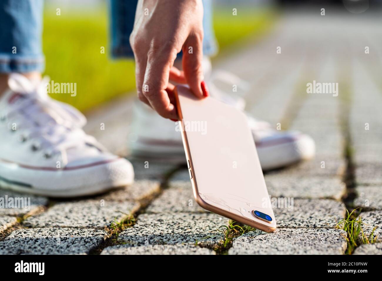 Donna mano che raccoglie telefono rotto da pavimentazione ciottolata dopo goccia, vista dal basso, primo piano. Smartphone con vetro rotto sul coperchio posteriore. Foto Stock