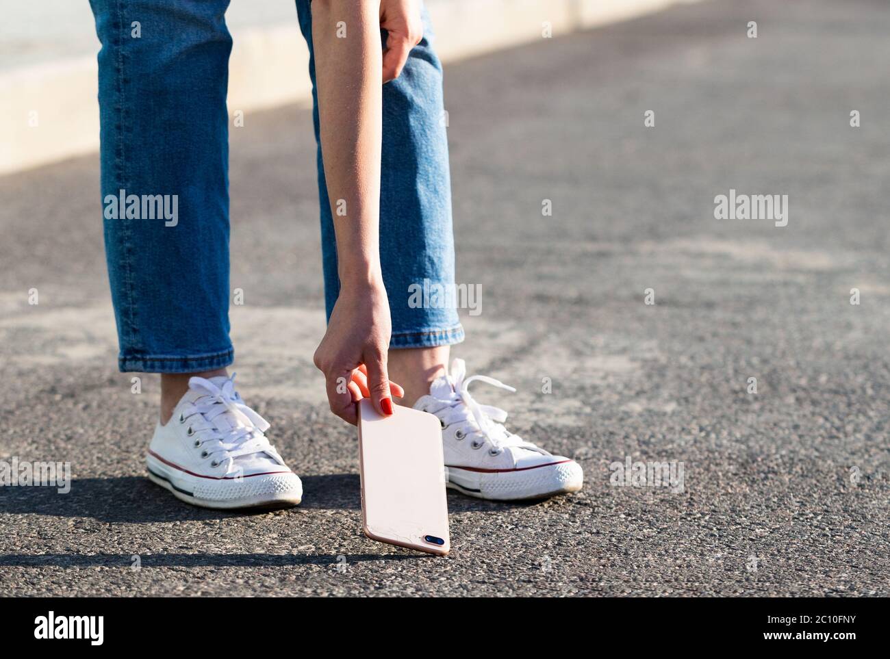 Donna mano che raccoglie telefono rotto da asfalto dopo goccia, vista dal basso, all'aperto. Smartphone con vetro rotto sul coperchio posteriore. Foto Stock