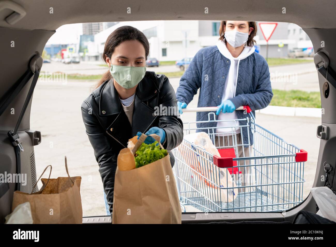 Giovane donna in maschera mettendo in macchina borse di carta piene di prodotti mentre il ragazzo si trova con il carrello dietro di lei Foto Stock
