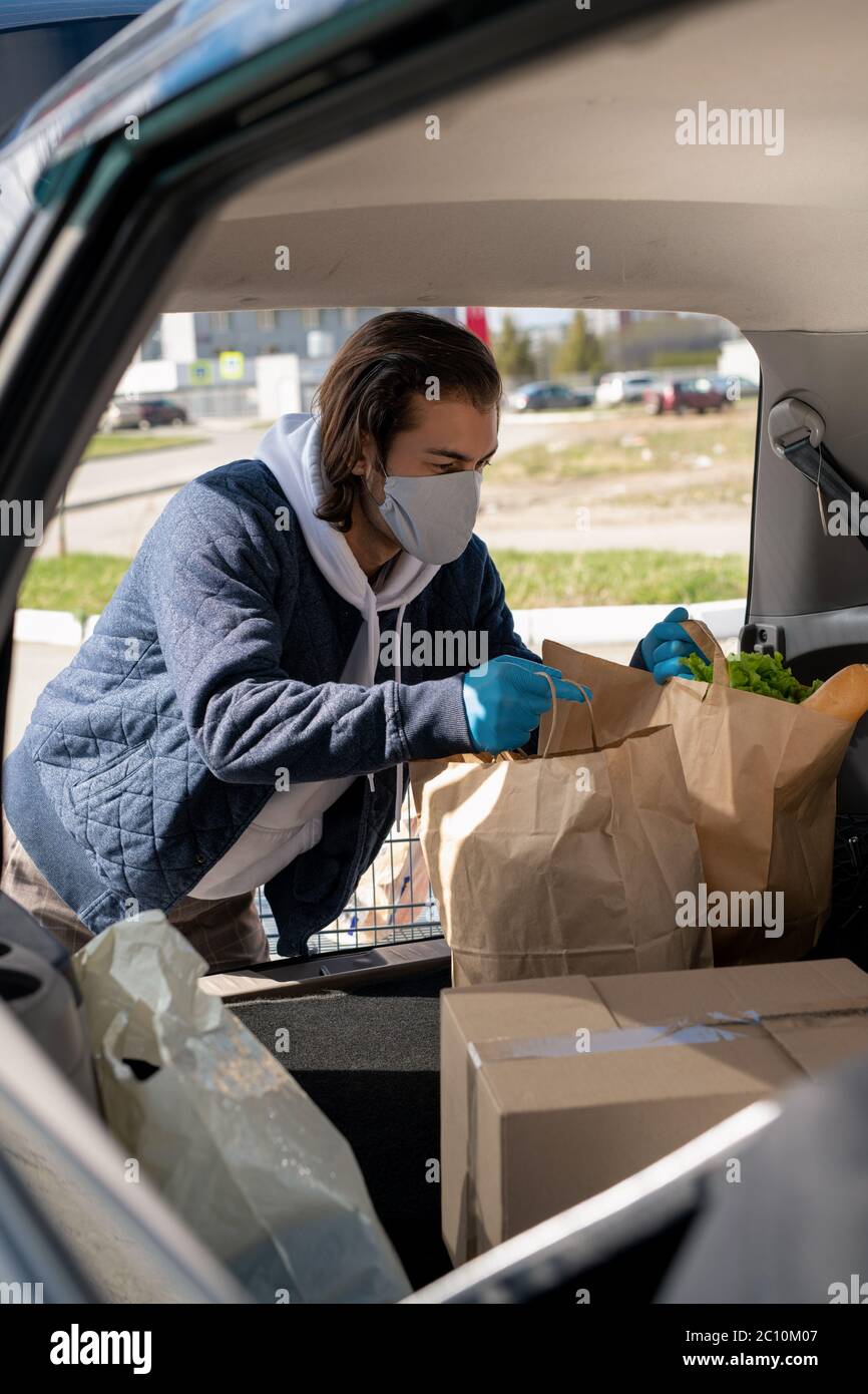 Uomo brunette in maschera e guanti che caricano i prodotti del supermercato nel bagagliaio dell'auto dopo lo shopping durante la pandemia Foto Stock