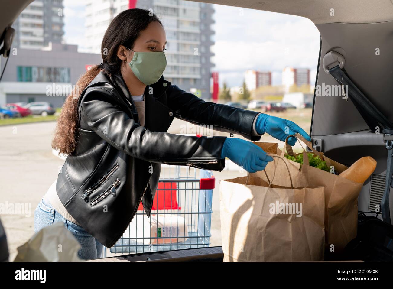 Giovane donna in maschera e guanti mettendo borse di carta nel bagagliaio dell'auto dopo lo shopping supermercato Foto Stock