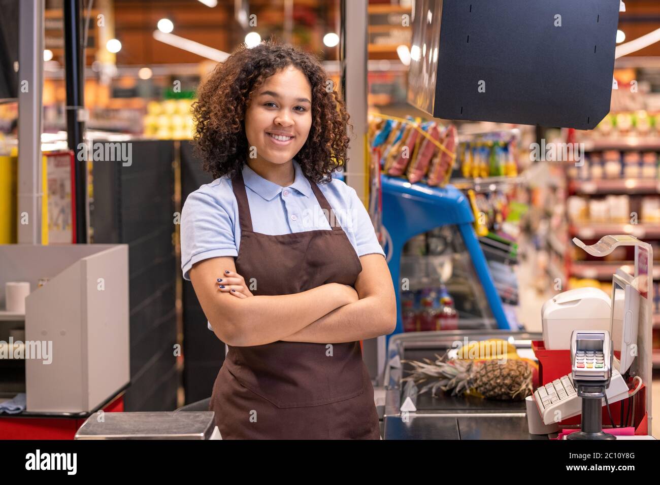 Bella femmina in grembiule in piedi da cashbox nel supermercato e incrociando le braccia da petto sullo sfondo di scaffali con prodotti alimentari Foto Stock