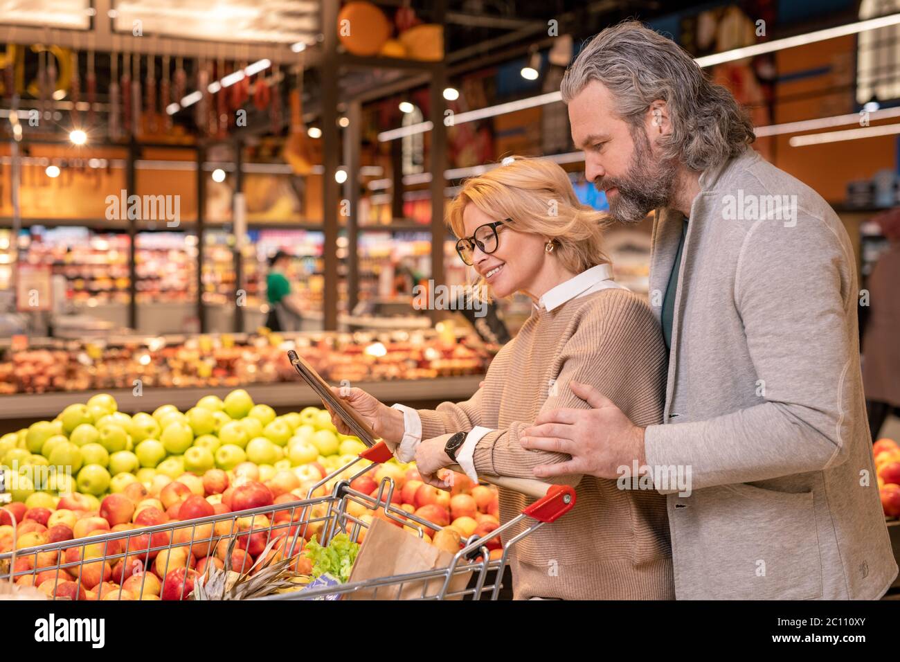 Felice giovane shopper donna che mostra la sua lista di acquisti marito in blocco note mentre entrambi camminando lungo esposizione con mele fresche Foto Stock