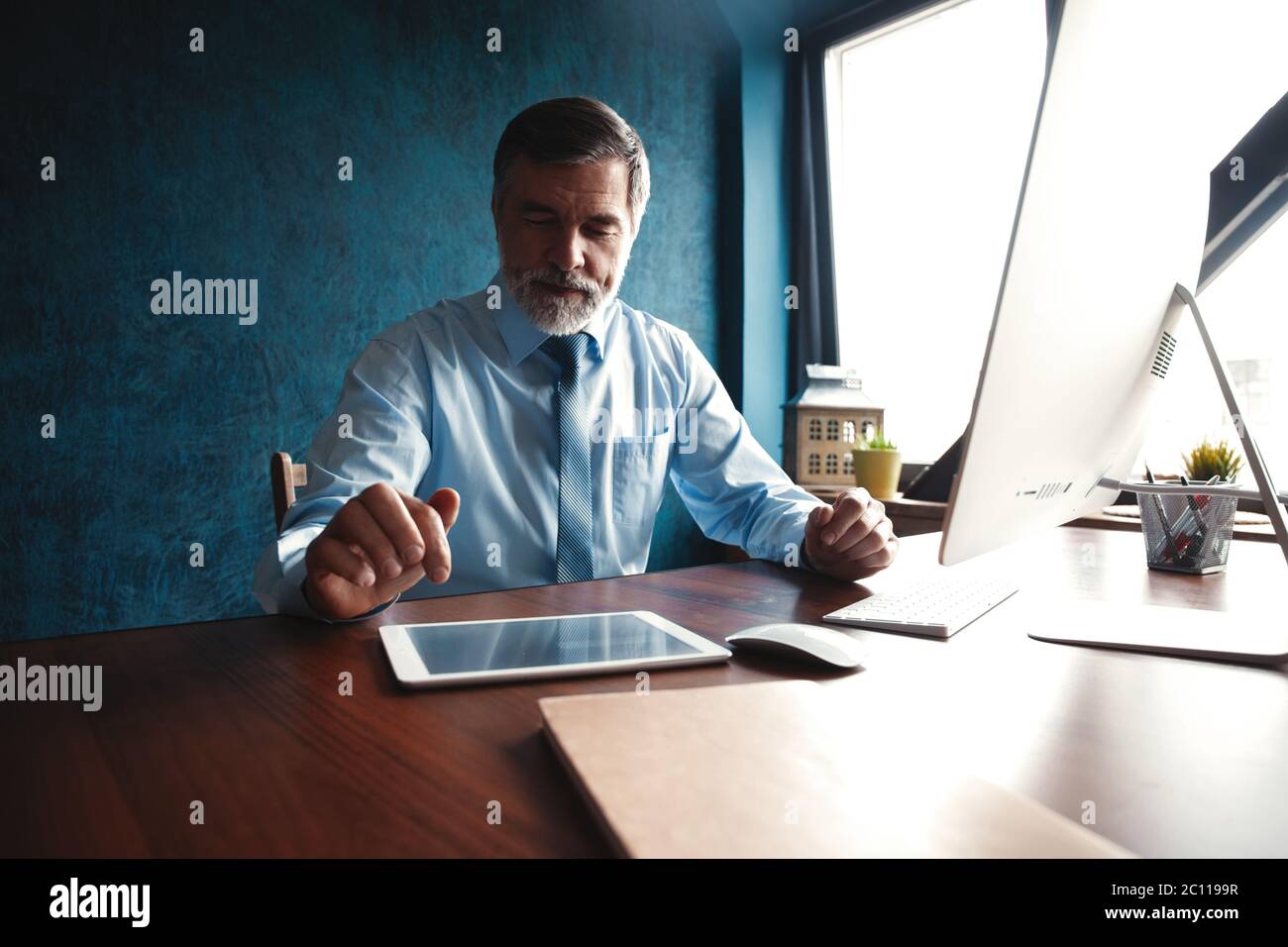 Consulente finanziario serio e maturo seduto al tavolo e controllando il rapporto finanziario utilizzando il tablet digitale in ufficio. Foto Stock