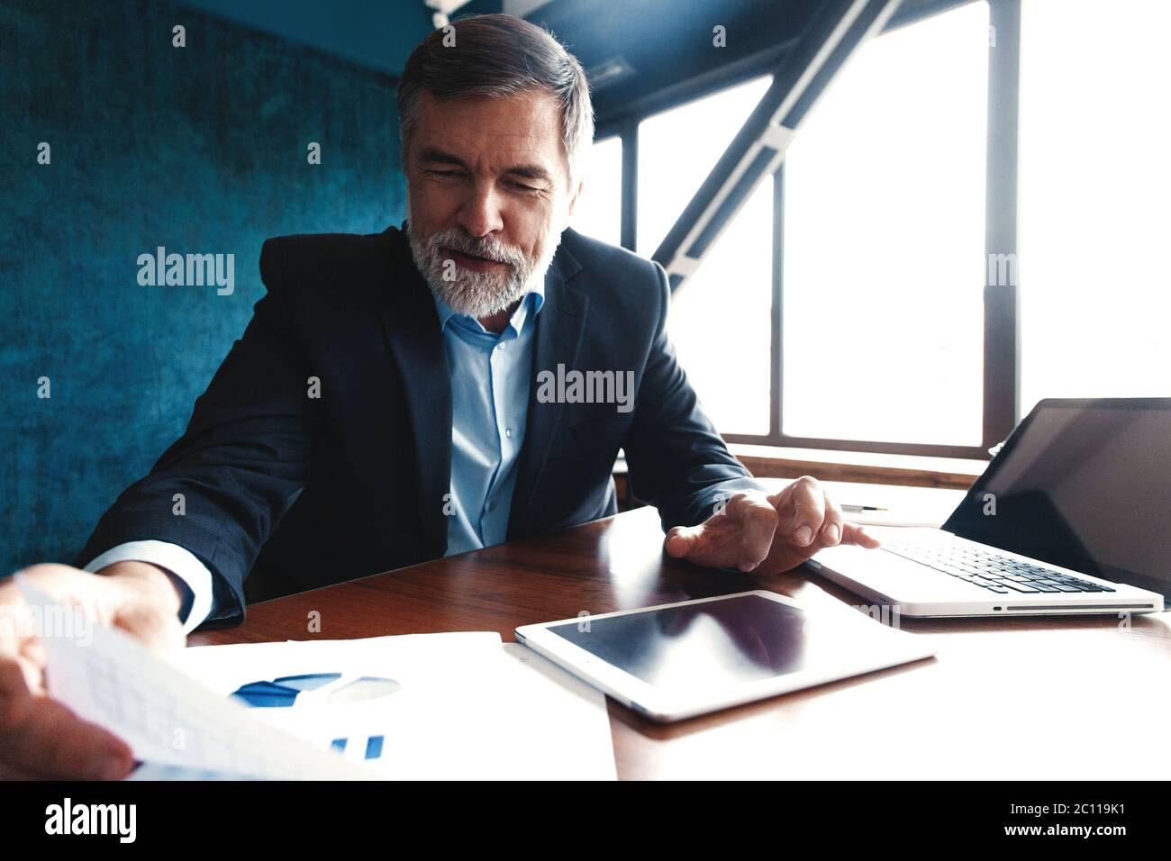 Consulente finanziario serio e maturo seduto al tavolo e controllando il rapporto finanziario utilizzando il tablet digitale in ufficio. Foto Stock