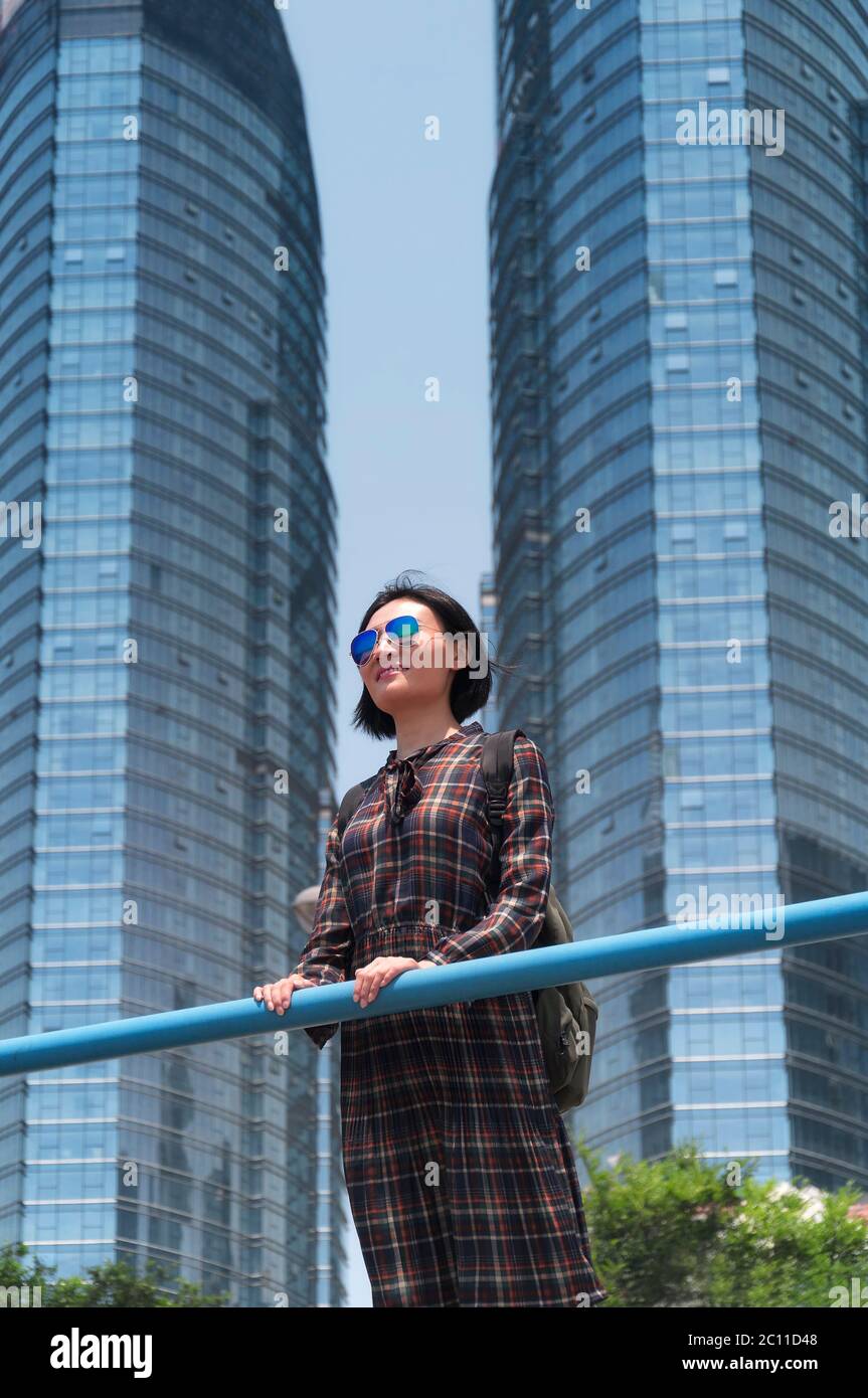 Una donna cinese felice appoggiata contro una ringhiera metallica con due grattacieli sullo sfondo che indossano un abito a plaid nella città di Qingdao, cina Foto Stock