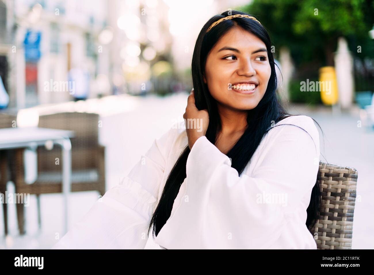 Giovane brunetta donna latina con capelli lunghi e abito bianco seduta sulla terrazza del bar. Stile di vita Foto Stock