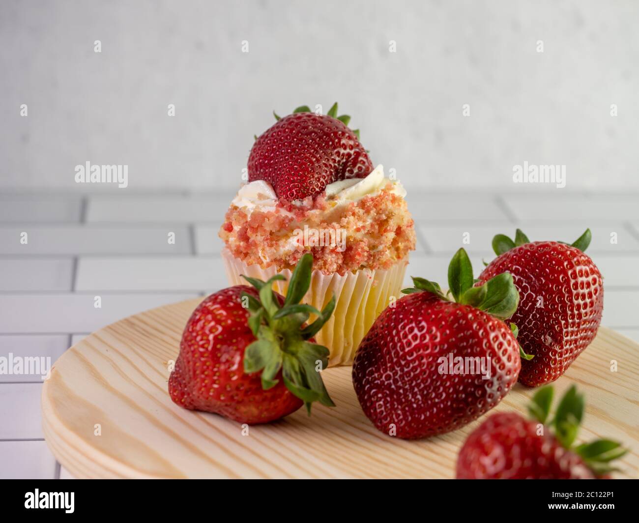 Fragole fresche su un tagliere rotondo in legno con una torta di fragole su un banco di piastrelle bianche della metropolitana con un muro di gesso dietro. Fre Foto Stock