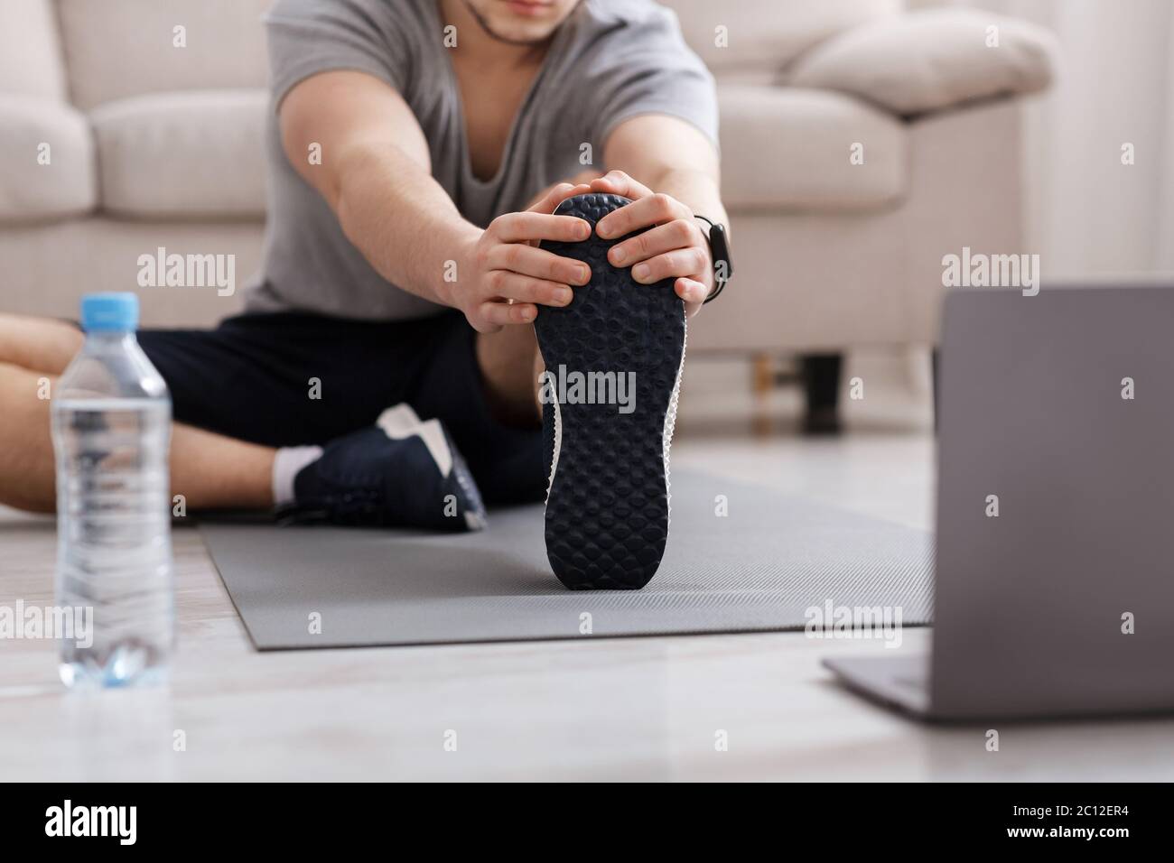 Palestra a casa. Uomo che fa stretching sul pavimento a casa, acqua e laptop vicino Foto Stock