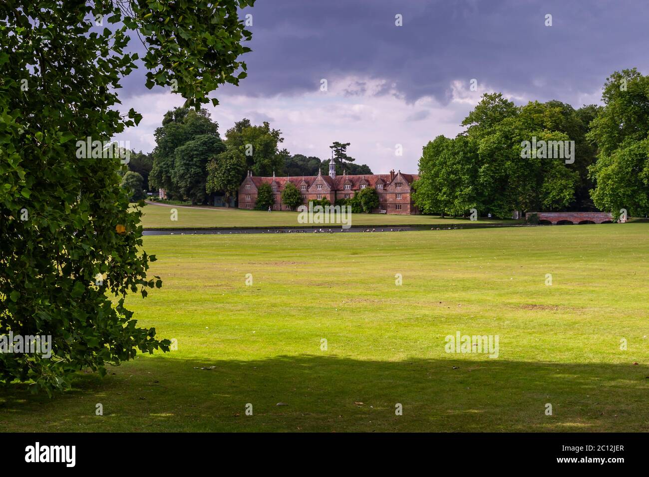 Il probabile blocco di stalle elisabettiano tardo della Audley End House si erge dietro un ampio prato vicino a Saffron Walden, Essex, Inghilterra, con un lago e uccelli acquatici in primo piano mentre nuvole scure di pioggia si accumulano sopra. Costruito in mattoni rossi, presenta una lanterna ottagonale con una guglia piombata all'incrocio della sua fila centrale, una caratteristica documentata per il blocco di stalle della tenuta della fine del XVI secolo. Thomas Howard, 1° conte di Suffolk, costruì la casa principale di Audley End stessa tra il 1605 e il 1614 in stile giacobino, una volta una delle più grandi case di campagna d'Inghilterra. English Heritage si è occupata della tenuta Foto Stock