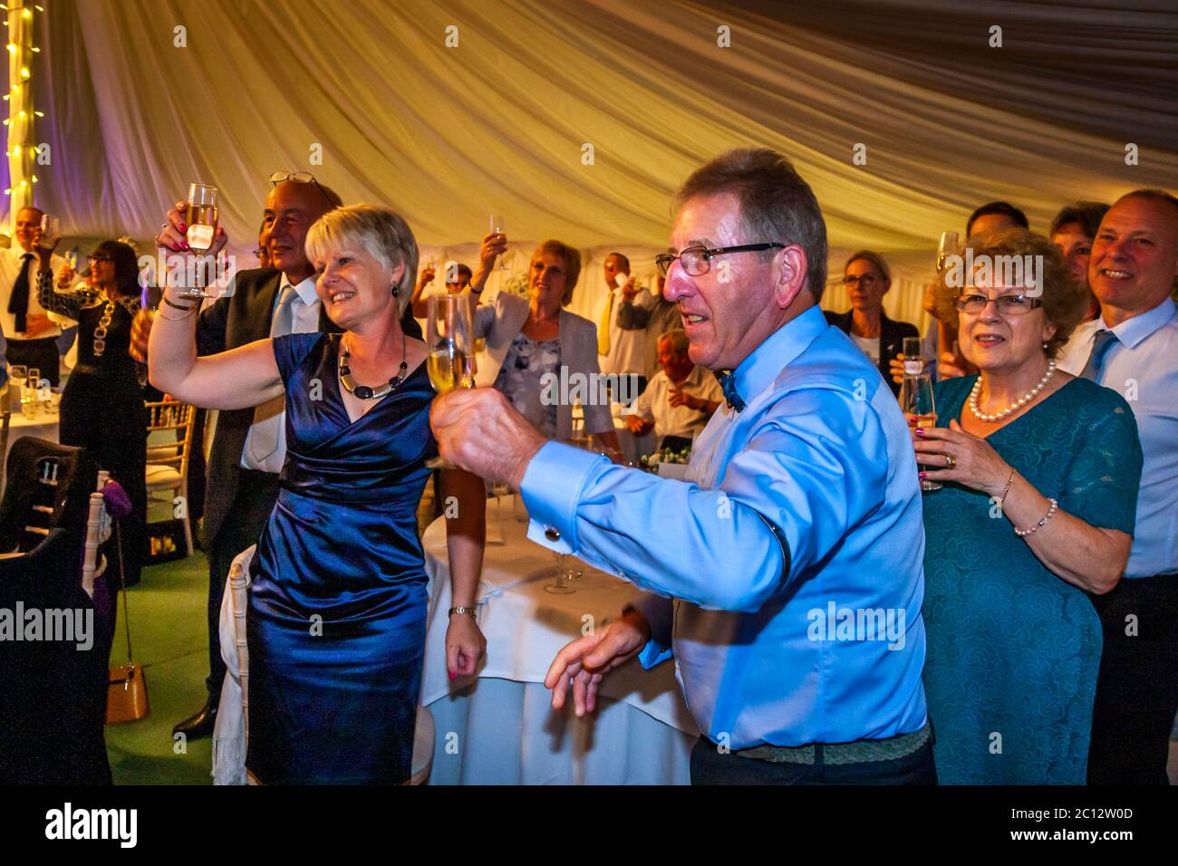La compagnia nuziale alza i calici di champagne per un brindisi all'interno della tenda festosamente illuminata durante la receptione di nozze a Longstowe Hall, South Cambridgeshire, Inghilterra. Foto Stock