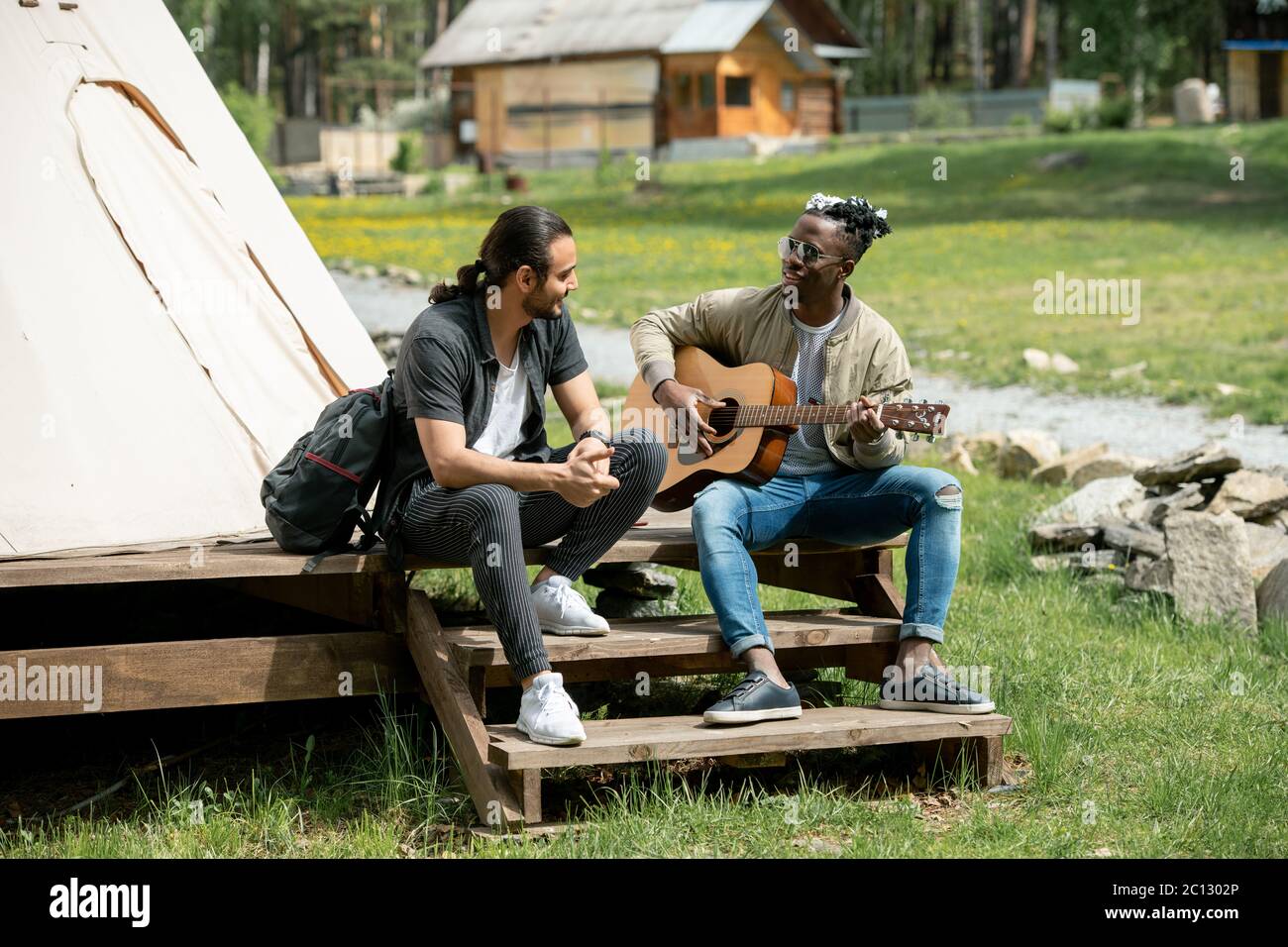 Ragazzo nero seduto sul gradino e suonare la chitarra per un amico mentre trascorrono il tempo al festival di campagna Foto Stock