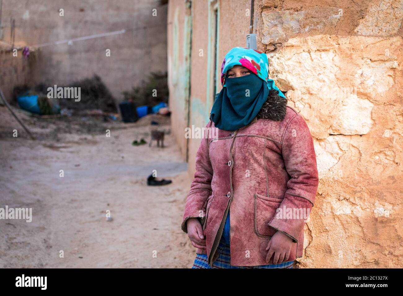 Ritratto di una donna nomade berbera con il viso coperto appoggiato su un muro di mattoni di fango in un semplice villaggio berbero nella provincia di midelt, marocco Foto Stock