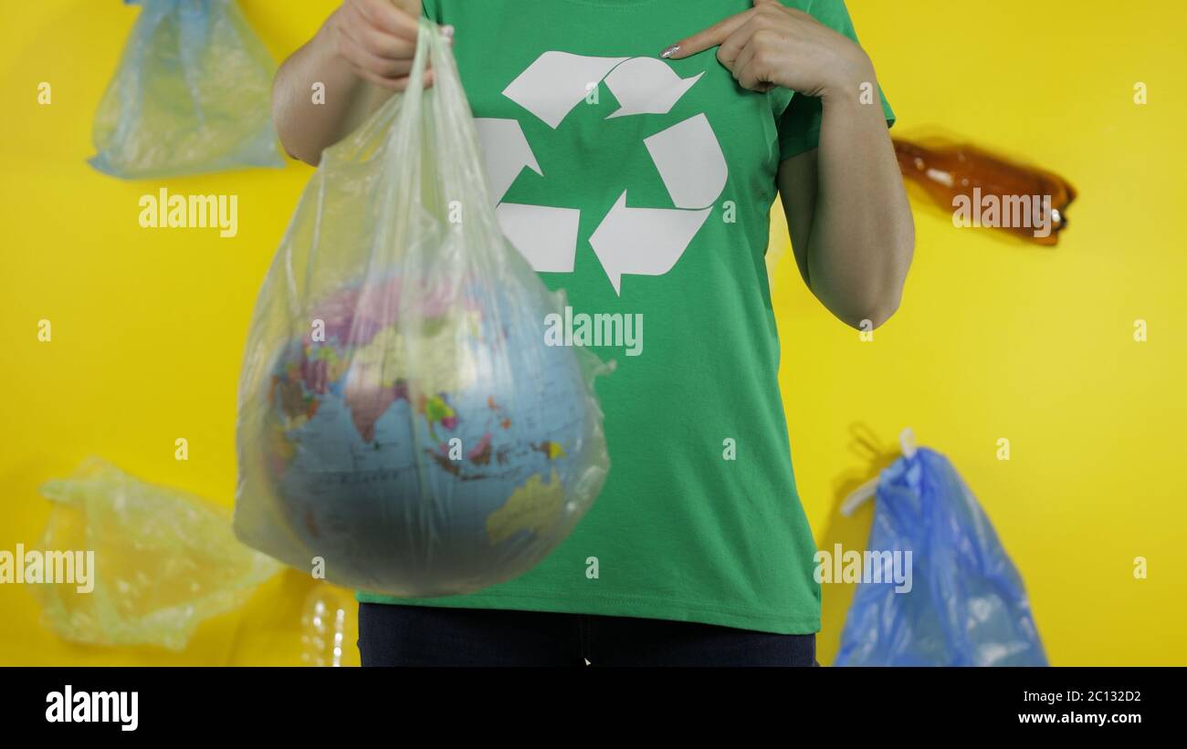 Irriconoscibile attivista femminile in t-shirt con logo riciclare con globo terrestre in pacchetto cellophane. Fondo giallo con sacchetti, bottiglie. Ambiente ecologico. Inquinamento della natura dei rifiuti di plastica Foto Stock