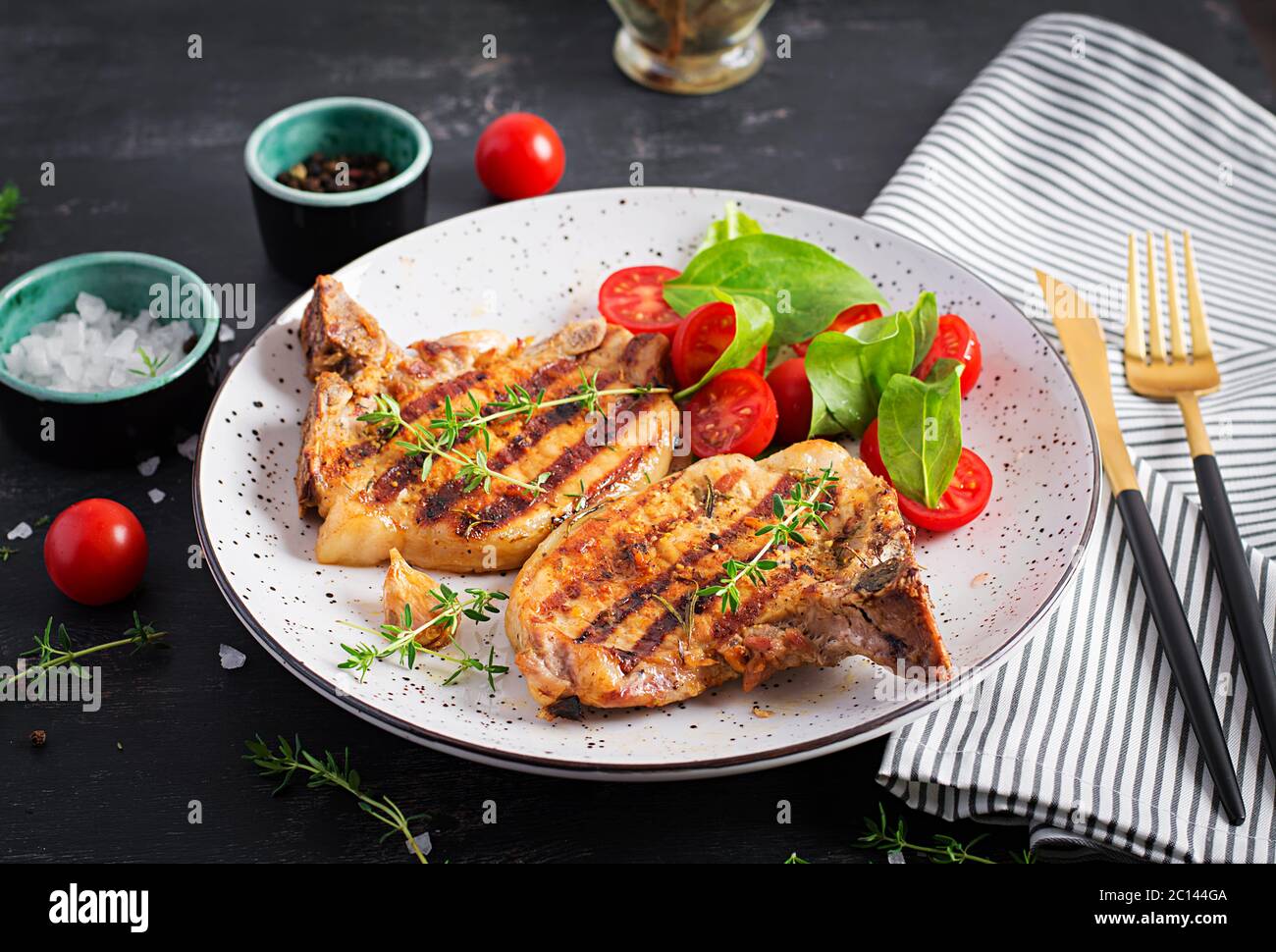 Bistecche di maiale alla griglia e insalata con pomodori in piatto su sfondo scuro. Foto Stock