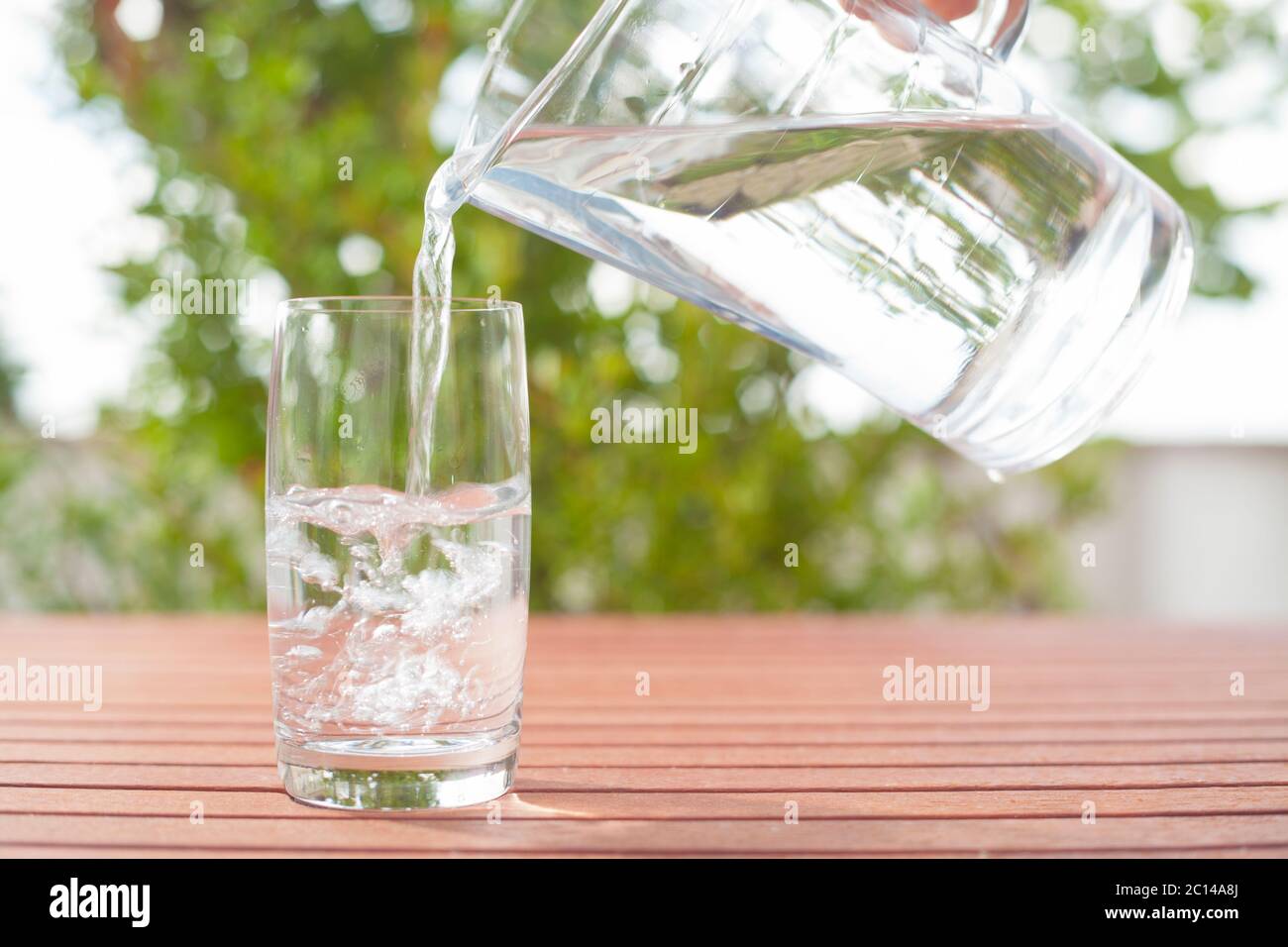 L'acqua potabile che scorre da una caraffa di vetro in un bicchiere – sfondo con piante Foto Stock