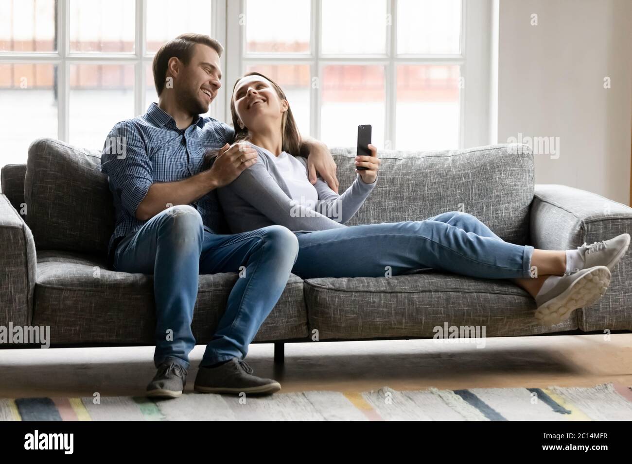 Felice giovane coppia abbracciando, rilassandosi sul divano, usando lo smartphone Foto Stock