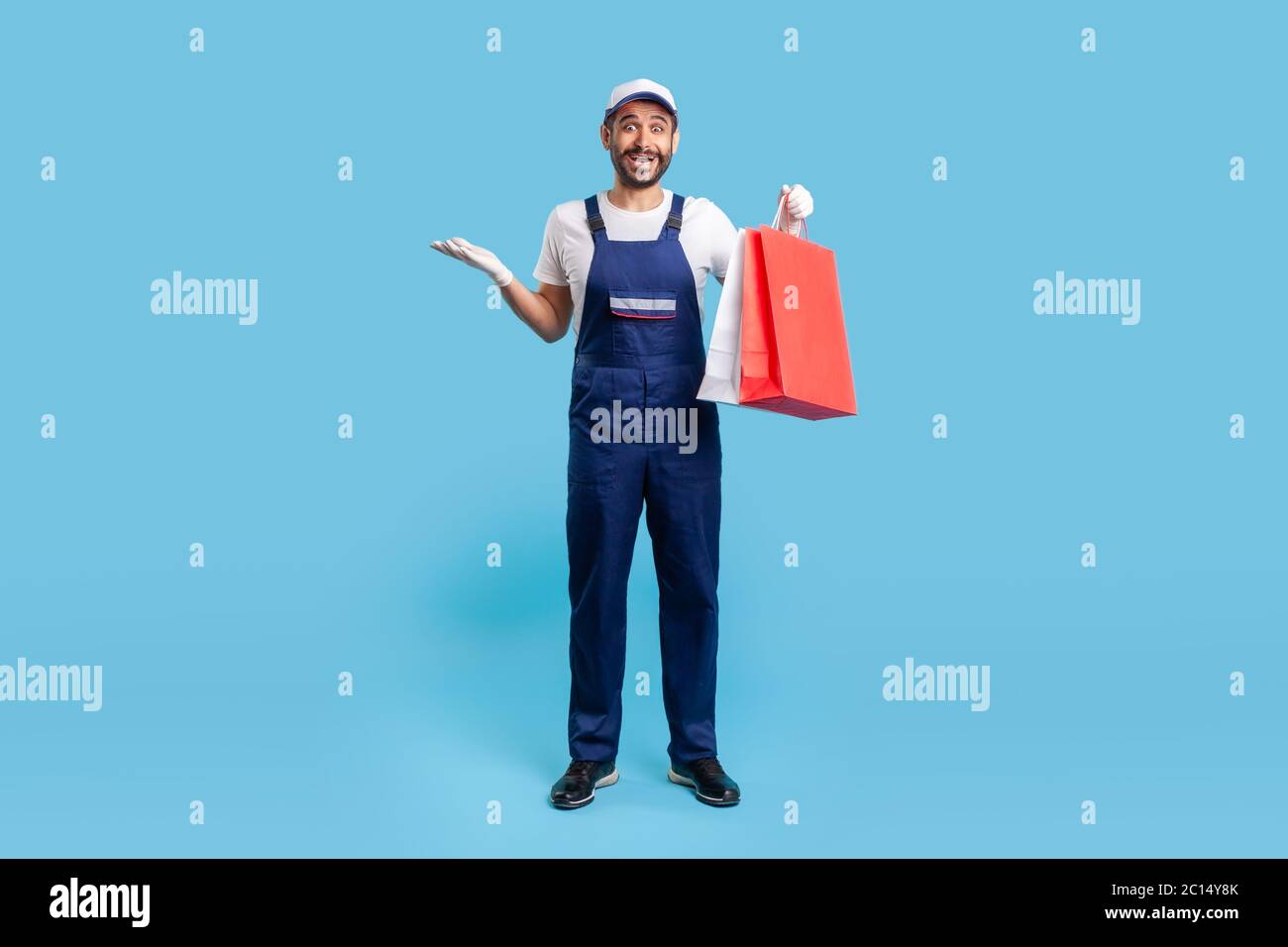 Ritratto di lunghezza completa del corriere sorpreso in uniforme che tiene le borse della spesa e che guarda scioccato, consegnando il pacco con le merci ordinate online alla moda Foto Stock