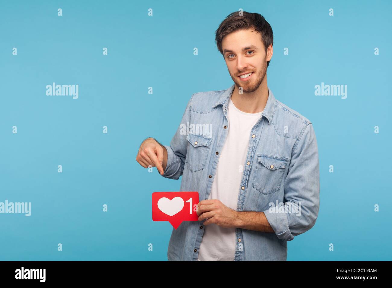 Clicca su mi piace e iscriviti! Ritratto di bel blogger uomo in camicia denim che punta al tasto cuore social media, contatore emoji, notifica seguace. Foto Stock