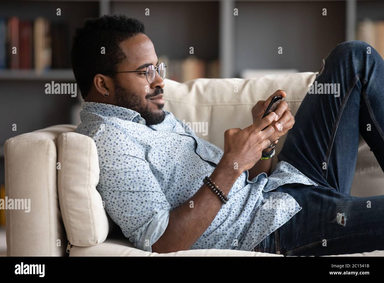 L'uomo africano giace sul divano che tiene lo smartphone leggere notizie sui media Foto Stock
