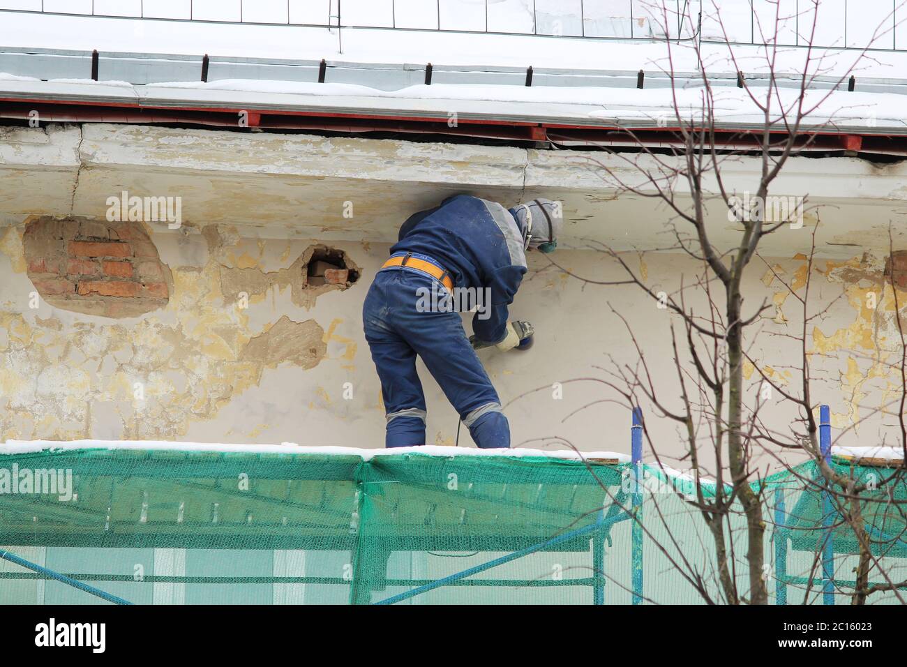 Il lavoratore in occhiali protettivi e un respiratore elabora la parete con una smerigliatrice angolare prima del restauro ed intonaci Foto Stock