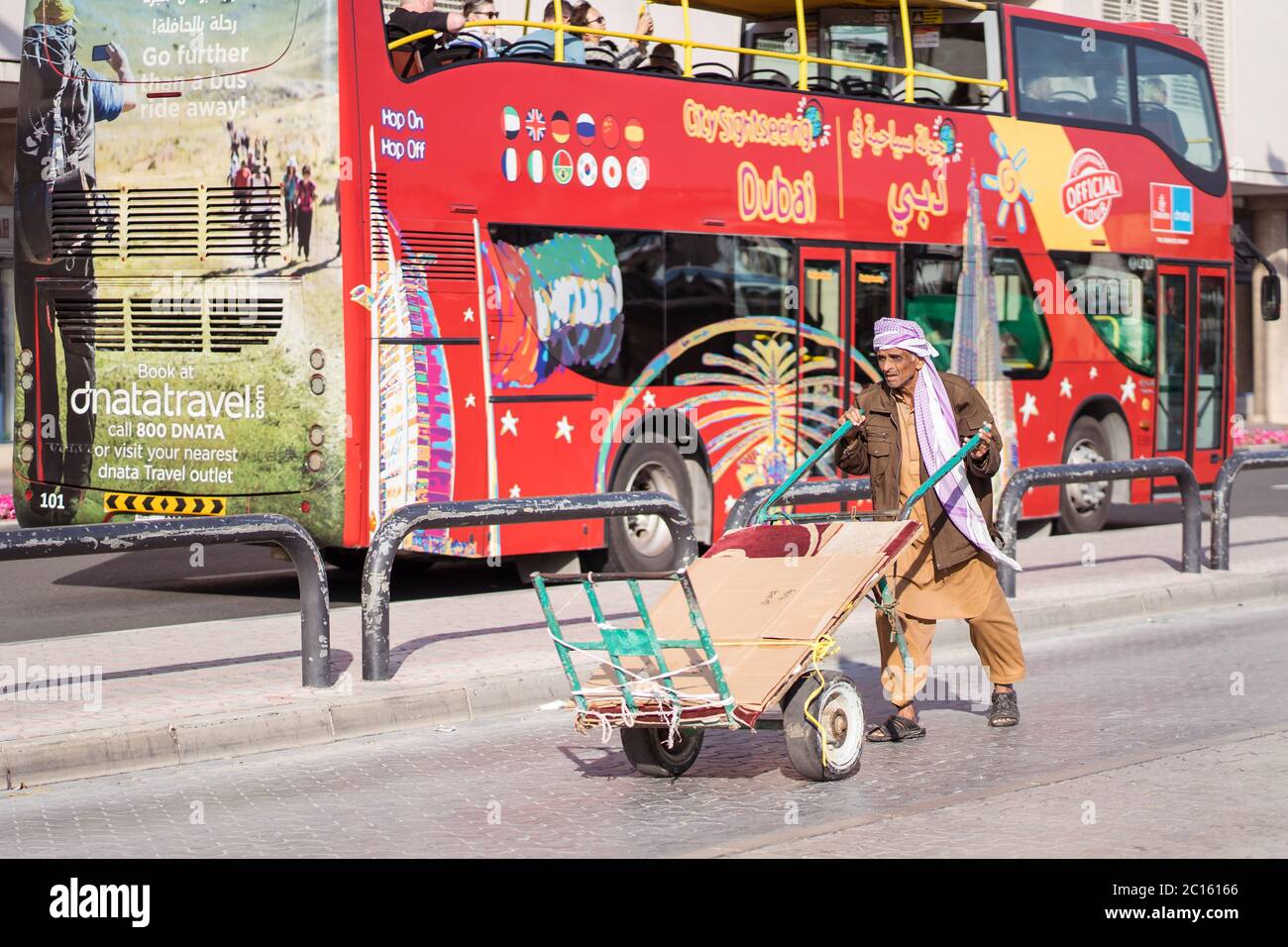 Dubai / Emirati Arabi Uniti - 1 febbraio 2020: Immigrato pakistano lavoratore che trasporta il carrello con il bus turistico rosso hop on hop off città sigthseeing dietro di lui Foto Stock
