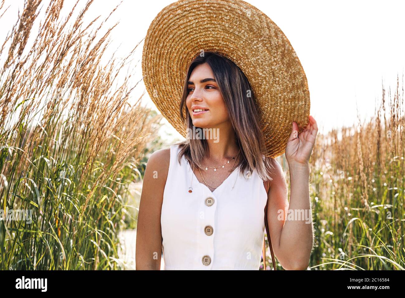 Giovane donna sorridente che indossa un cappello di paglia e cammina nel parco Foto Stock