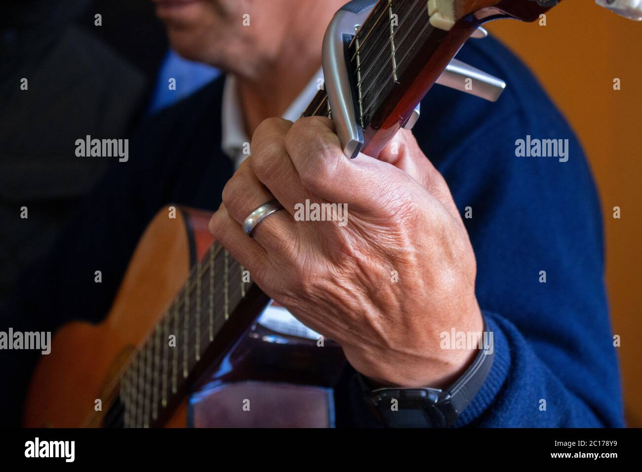 Avvicinati alla mano di un anziano seduto tenendo una chitarra con un anello di nozze sul dito su uno sfondo marrone Foto Stock
