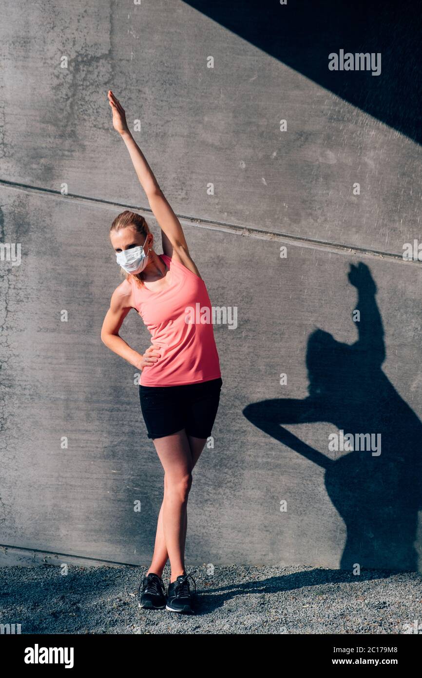 ritratto verticale di sana donna caucasica in abbigliamento sportivo con maschera protettiva che si estende all'aperto al tramonto su un fondo di parete in cemento Foto Stock