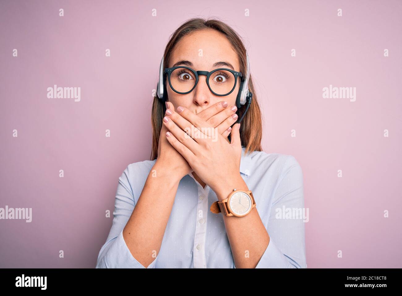 Giovane bella agente del call center donna che indossa occhiali che lavorano con cuffie scioccato coprendo la bocca con le mani per errore. Concetto segreto. Foto Stock