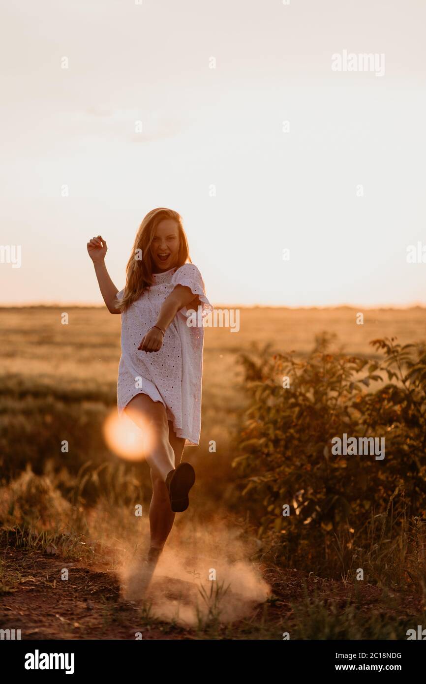 Giovane bella donna che fa polvere con il piede e urla nel campo di arancio di grano in una giornata estiva soleggiata. Impazzire. Sentendosi liberi e felici Foto Stock