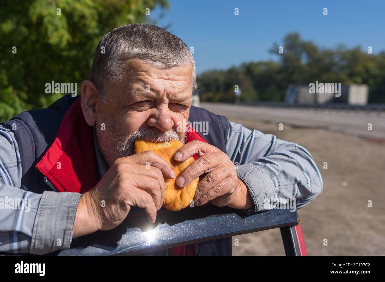 Ritratto di un autista anziano affamato che mangia patti appoggiando i gomiti sulla porta della sua auto Foto Stock