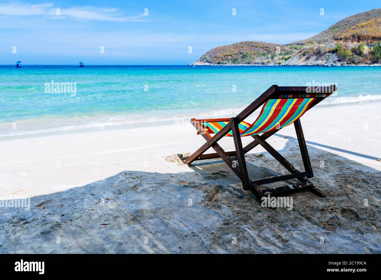 Una sedia a sdraio si trova all'ombra sulle rive di una spiaggia di sabbia sul mare. Il colore bruoso dell'acqua e del cielo blu con nuvole rotte nella b Foto Stock
