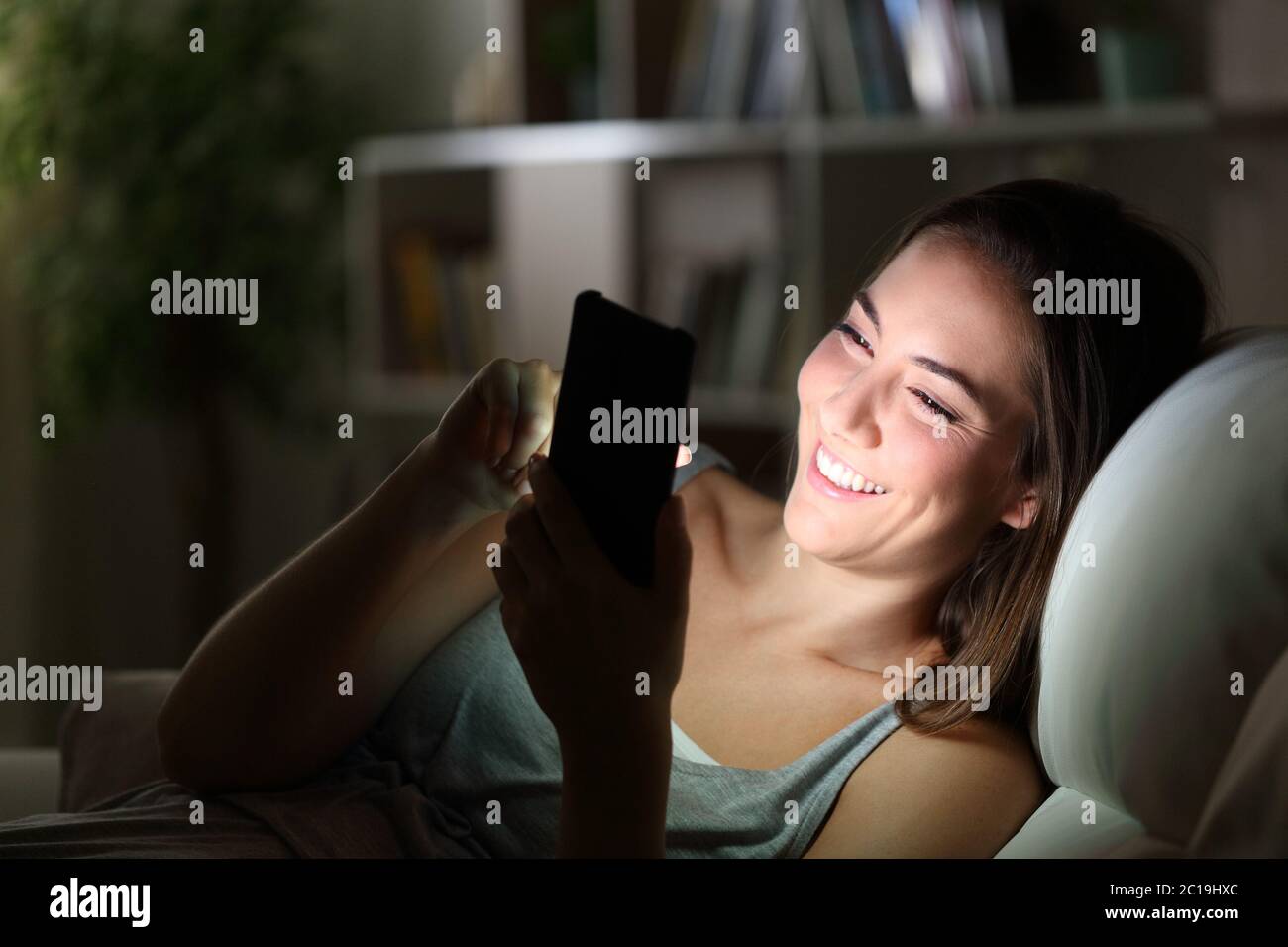 Donna felice che usa il telefono con il viso illuminato seduto su un divano a casa di notte Foto Stock