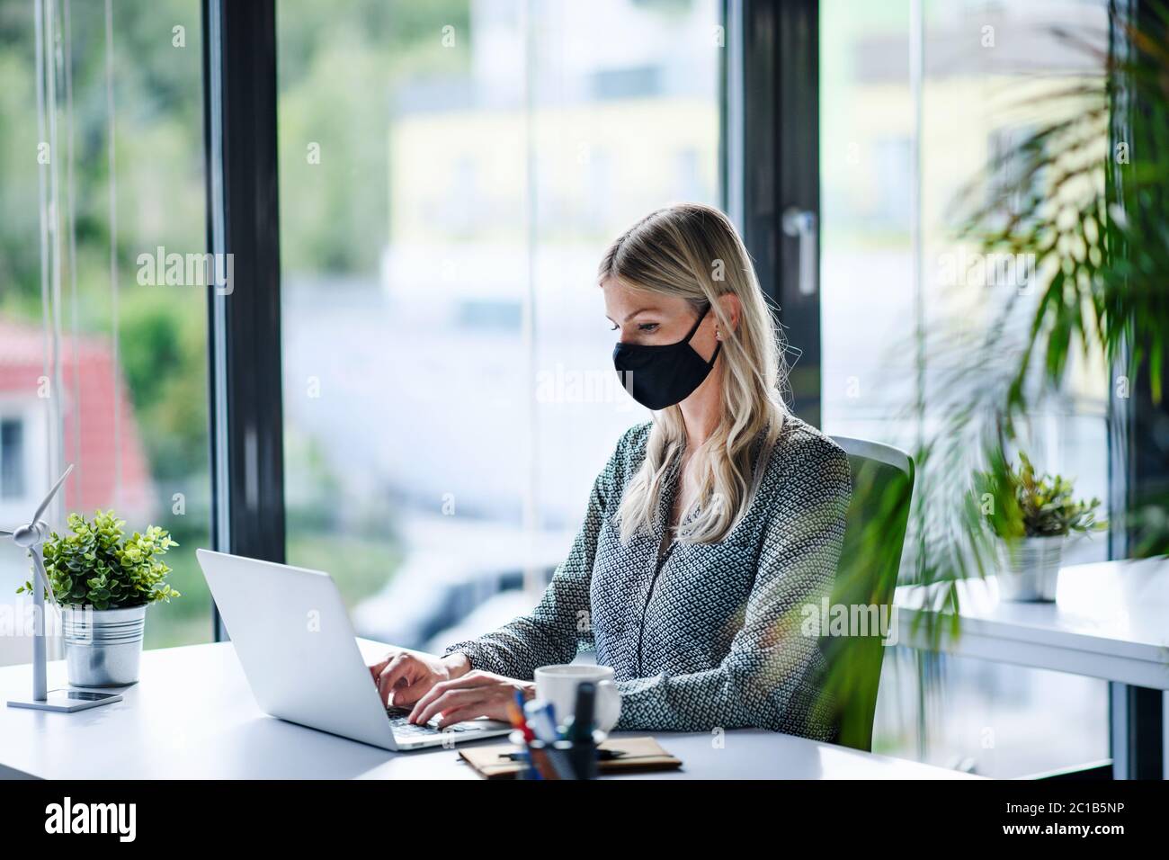 Giovane donna con maschera facciale di nuovo al lavoro in ufficio dopo il blocco, utilizzando il laptop. Foto Stock