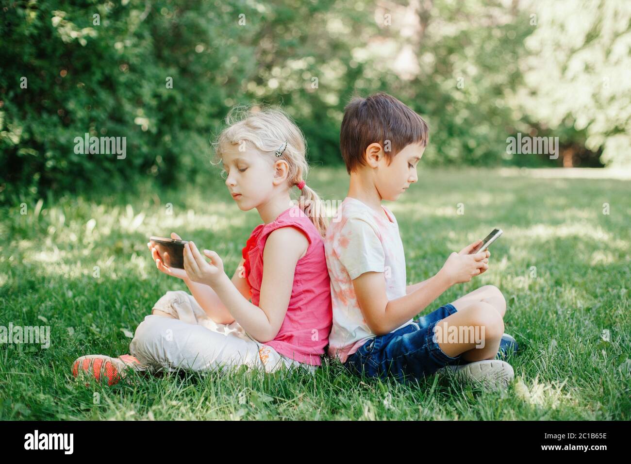 Ragazzo e ragazza giocano con gli smartphone all'aperto. Dipendenza dallo schermo dei gadget digitali per bambini. Bambini amici che giocano online seduti sull'erba nel parco. Foto Stock