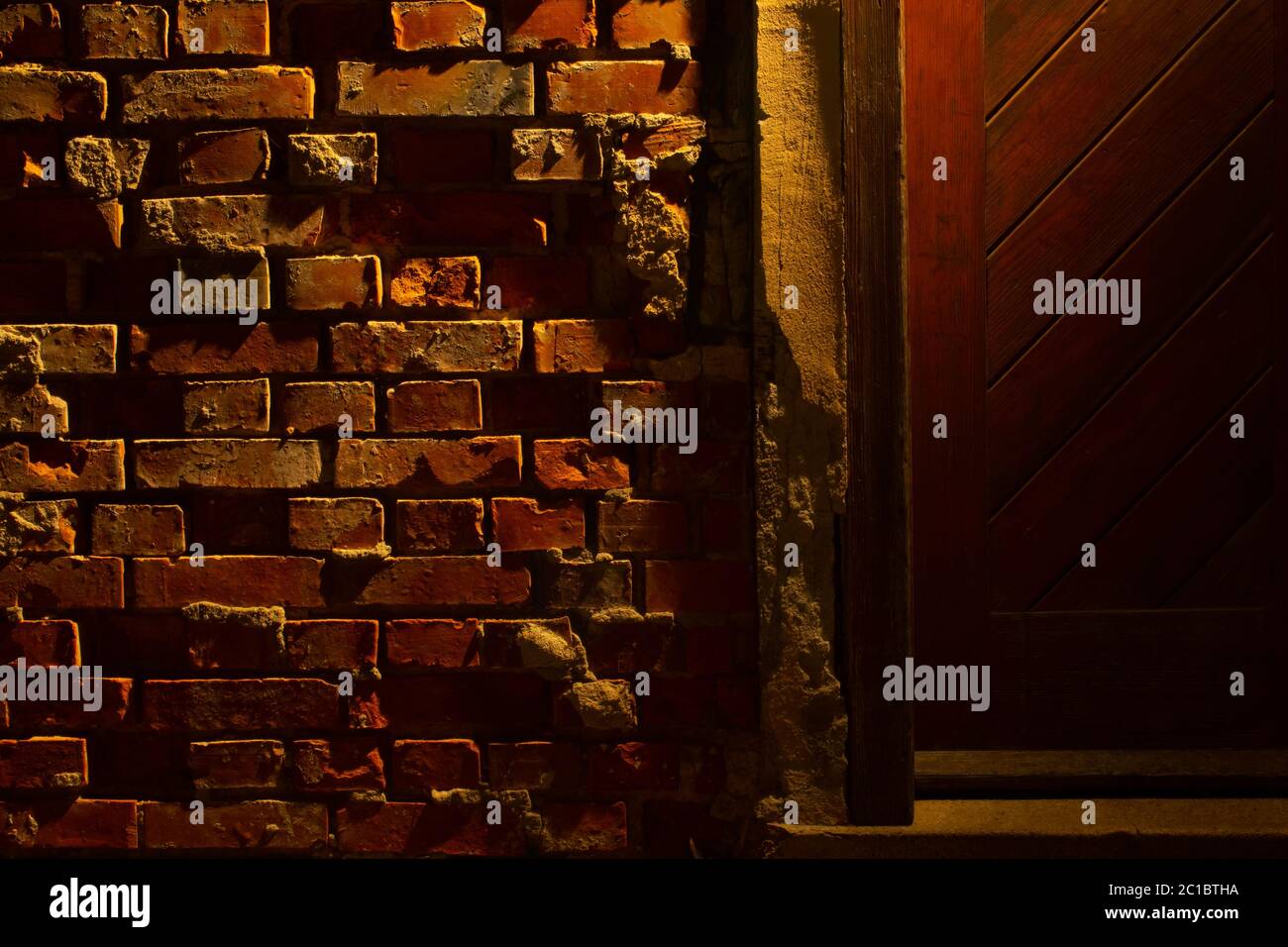 Vecchio grugnoso e danneggiato muro di mattoni rossi di notte accanto ad una porta di legno con luce di una lanterna che splende su di essa Foto Stock