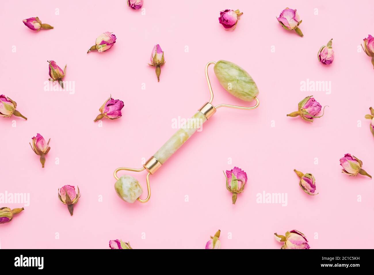 Rullo massaggiante per viso con due pietre di giada su sfondo rosa con fiori di rose essiccate intorno. Concetto di assistenza sanitaria. Vista dall'alto Foto Stock
