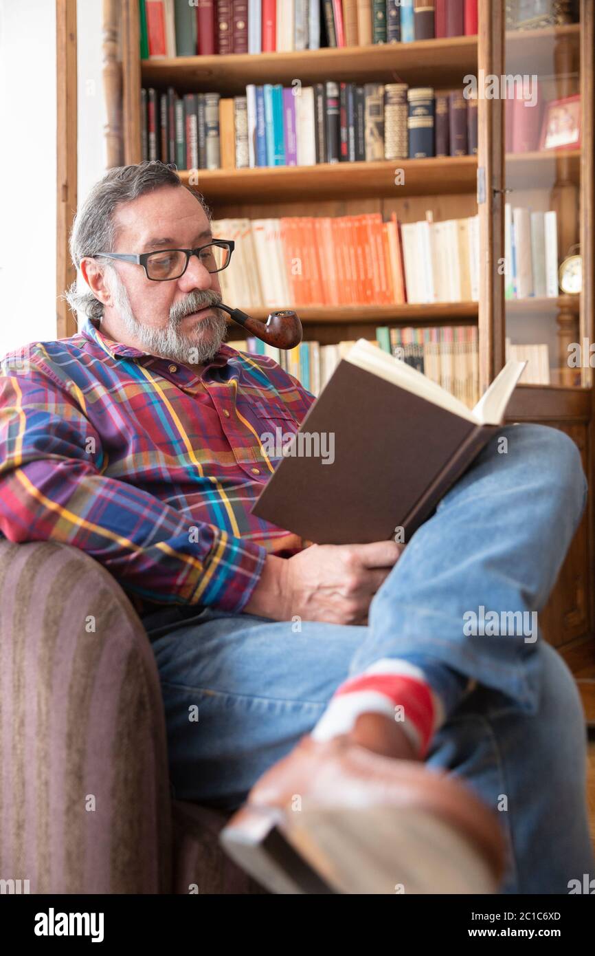 Ritratto di un uomo anziano che legge un libro accanto alla sua biblioteca mentre fuma un tubo. Concetto di soggiorno a casa. Foto Stock