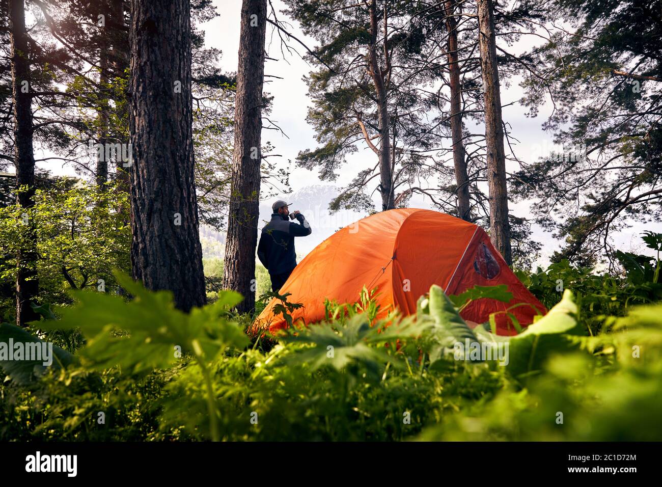 Il turista bearded sta bevendo il caffè vicino alla tenda arancione nella foresta lussureggiante alle montagne Foto Stock