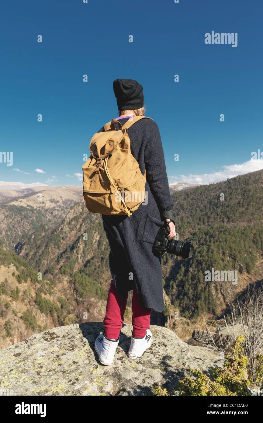 Una fotografa di viaggio hipster ragazza in un cappello e con uno zaino in piedi su una roccia e guardando le colline. Foto Stock