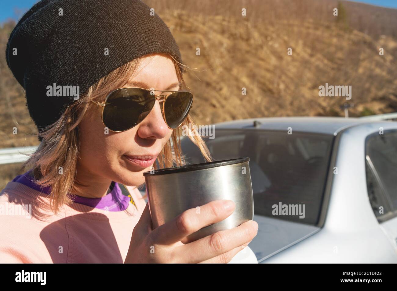 Una ragazza hipster in occhiali da sole e un cappello nero beve tè o caffè da una tazza da un thermos nella natura in un viaggio contro t Foto Stock