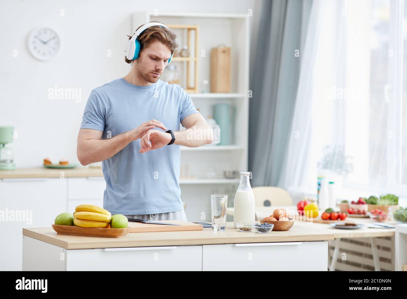 Giovane uomo che indossa le cuffie e controlla il tempo sull'orologio mentre cucina la colazione in cucina Foto Stock
