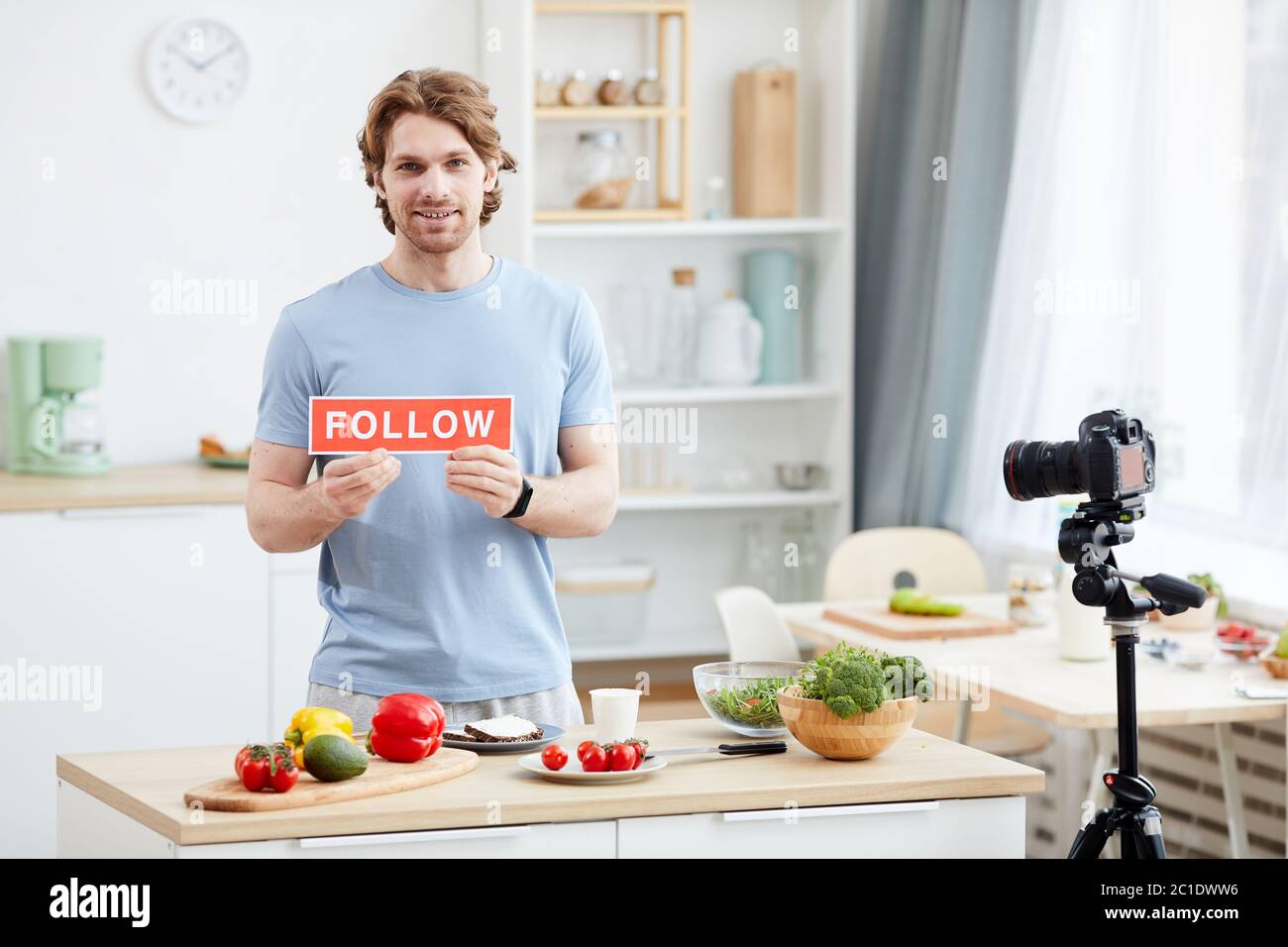 Ritratto di giovane uomo con placard seguire sorridente a macchina fotografica e sparare blog per i suoi seguaci in cucina domestica Foto Stock